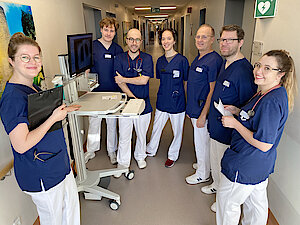 Männer und Frauen in blauen Kassaks und weißen Hosen stehen um einen mobilen PC-Arbeitsplatz und schauen in die Kamera.