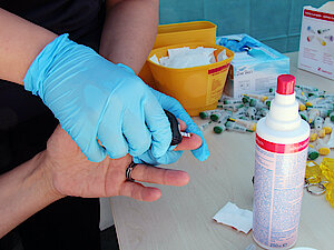 Quartiersfest_8947 Eine behandschuhte Hand führt einen Blutzuckertest am Finger durch, wobei Desinfektionsmittel und medizinisches Material auf einem Tisch liegen.