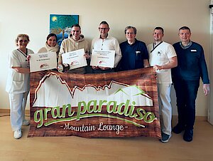Männer und Frauen, einige von ihnen in weißen und dunkelblauen Kassaks, andere in normaler Kleidung, stehen vor einer Wand und halten Zettel sowie ein Banner mit der Aufschrift "gran paradiso" in der Hand.
