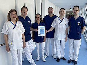 260324_Rezertifizierung ATZ Männer und Frauen in medizinischer Dienstkleidung stehen auf einem Flur und halten ein Zertifikat in den Händen.