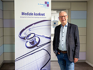 Ein Mann in Hemd und Sakko steht vor einem Roll-Up. Darauf steht "Medizin konkret".